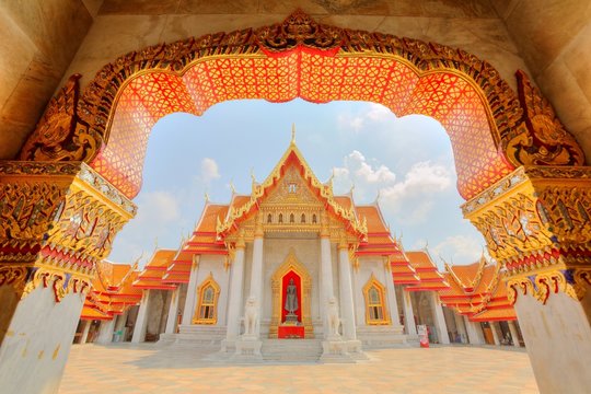 View through the gate to Wat Benchamabophit or The Marble Temple in Bangkok City, Thailand ~ Beautiful scenery of a colorful Buddhist temple with a decorated gate and golden roofs under sunny sky