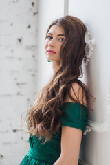 beautiful brunette girl with long hair in green dress standing near the white wall looking at the camera close-up of copyspace