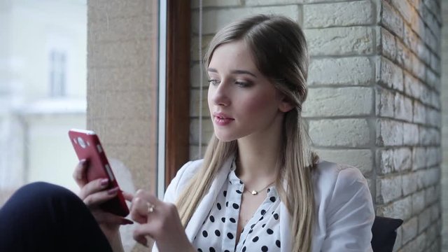Young beautiful blonde start-upper sits in the modern office, texting on her mobile phone then looks to the camera and smiles.