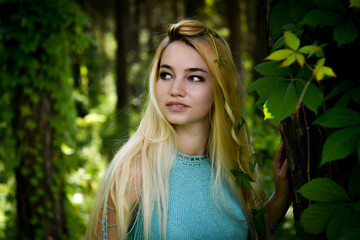 Pretty young blonde girl with long hair in turquoise dress standing in the green forest where trees are enlaced with lianas