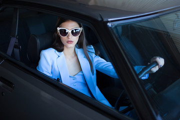Fashion girl driving a car in a blue suit. Stylish woman sitting in the car and holding steering wheel. Shot through the side window.