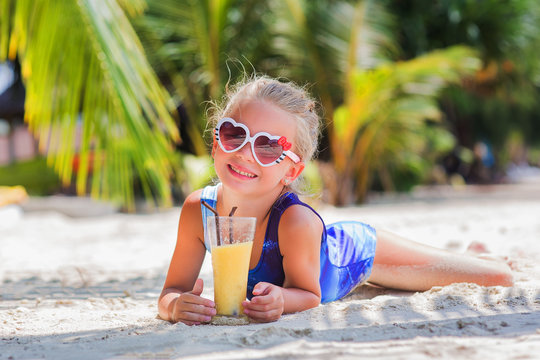 little cute girl on the sand at the beach in sun glasses with a glass of exotic cocktail juice
