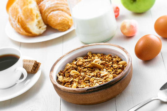 Healthy food for breakfast on a white wooden table. Oatmeal granola with nuts, coffee, croissants, eggs, milk and fruits for delicious healthy meal.