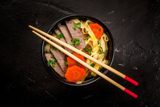 Beef soup with noodles and carrots in the Asian style. With chopsticks, on the black concrete table, copy space top view
