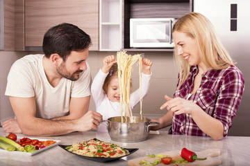 Young couple with kid having fun in kitchen while preparing spaghetti and salad