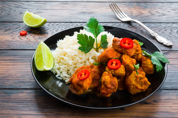 Traditional Indian dish, chicken curry with fresh chilli pepper, lime and parsley and boiled rice on a black ceramic plate on the wooden table.