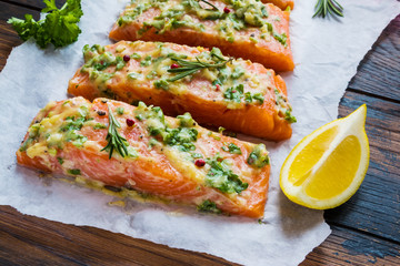 A process of cooking, pieces of raw salmon have been coated by garlic herb butter mixture on parchment paper, wooden table.