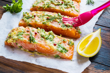 A process of cooking, pieces of raw salmon have been coated by garlic herb butter mixture on parchment paper, wooden table.