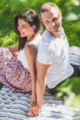 Fototapeta premium Vertical top view of a attractive couple in love sitting on a picnic blanket. Caucasian happy dreamy man and smiling brunette woman looking away on plaid on green grass.