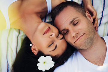 Top view of a beautiful horizontal portrait of a couple in love lying on a picnic blanket. Happy caucasian man and woman with white flower in hair with closed eyes on plaid in tropical.  