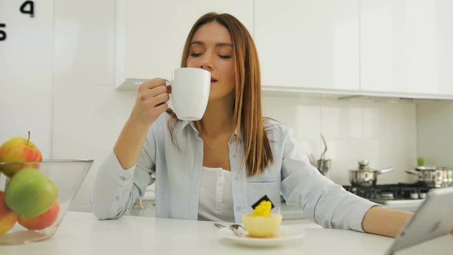 Pretty young woman drinking coffee watching her tablet computer and scrolling.