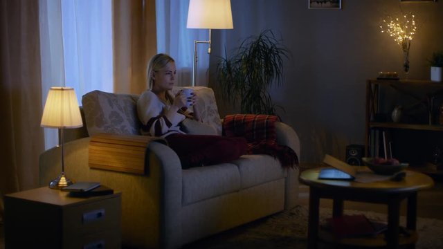Beautiful Woman Sits on a Couch in Her Cozy Living Room. She Watches TV and Drinks Hot Drink. Shot on RED EPIC-W 8K Helium Cinema Camera.