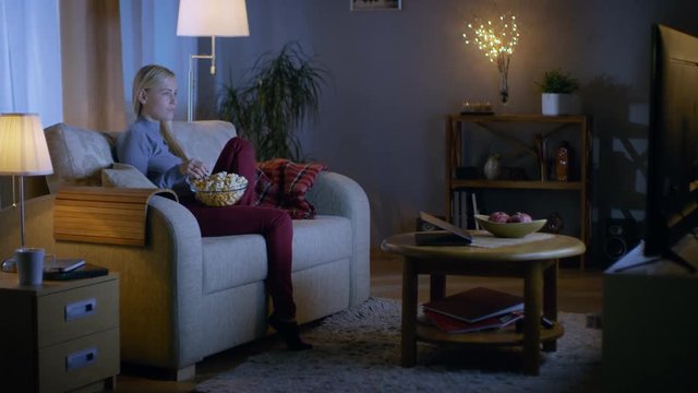 Beautiful Young Woman Sitting On Her Sofa in a Living Room. She eats Popcorn and Watches Television. It's Evening, Room Looks Cozy. Shot on RED EPIC-W 8K Helium Cinema Camera.