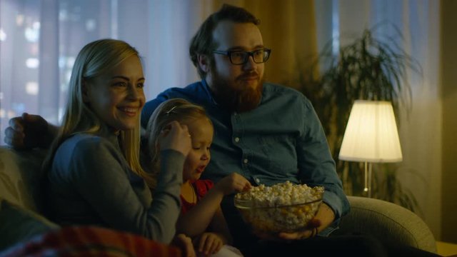 Father, Mother and Little Girl Watching TV. They Sit on a Sofa in Their Cozy Living Room and Eat Popcorn. It's Evening. Shot on RED EPIC-W 8K Helium Cinema Camera.