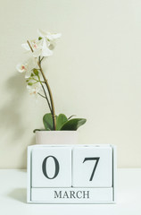 Closeup white wooden calendar with black 7 march word with white orchid flower on white wood desk and cream color wallpaper in room textured background , selective focus at the calendar
