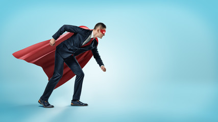 A businessman in a superhero red cape and a mask standing in starting line position on blue...