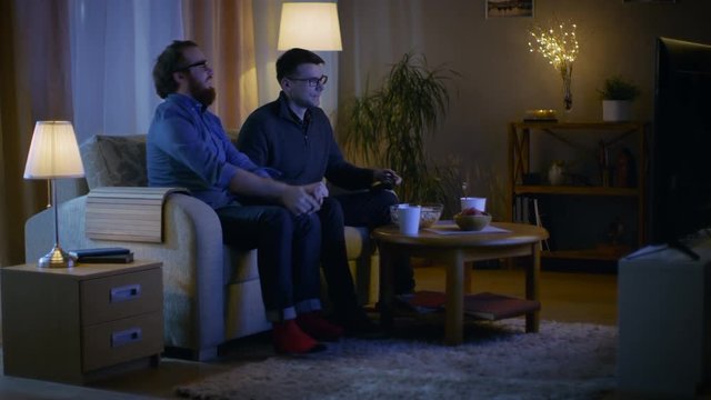 In the Evening Two Friends are Sitting on a Sofa in the Living Room and  Playing Competitive Video Games. One of Them Wins and He is Enjoying His Success. Shot on RED EPIC-W 8K Helium Cinema Camera.