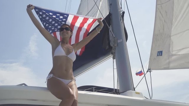 Beautiful young woman in white swimsuit  is holding an american flag on a yacht's deck