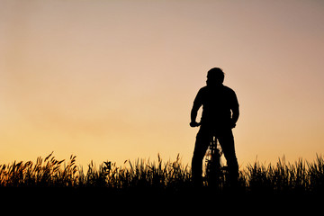 Silhouette of man with bicycle on grass field at the sky sunset, color of vintage tone and soft focus concept journey in holiday