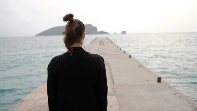 Shooting from back woman walking on sea shore looking into distance. Dreaming female in ripped jeans, black jacket, brown sweater and boots walks on beach, goes to the end of pier enjoying nature