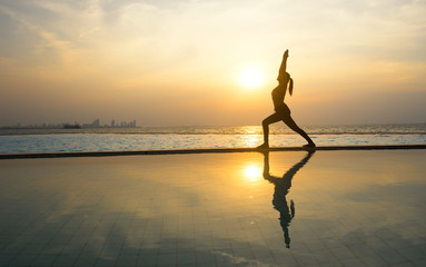 Silhouette young woman practicing yoga on swimming pool and the beach at sunset..