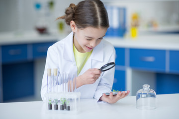 Smiling girl scientist holding magnifier and looking at green plant in soil