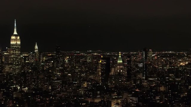 AERIAL HELI SHOT: Flying above Midtown Manhattan skyline, luxury residential buildings, condominiums and apartments lit up with lights at night towards glowing city lights in Brooklyn neighborhood