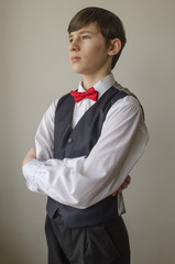 Teenager boy in a white shirt and a waistcoat with the red bow tie on a light background
