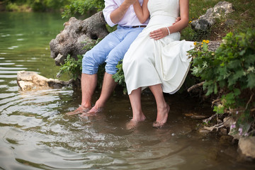 The bride and groom sitting on the shore of the lake. Foot splashing in the water.