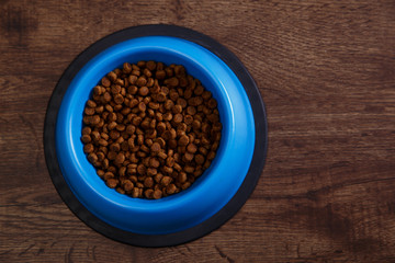 Bowl of dry kibble dog food. Healthy pets feed. Blue plate on wooden rustic background.