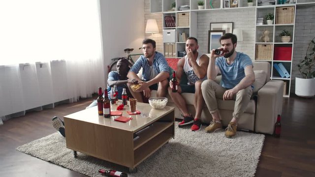 Slow motion shot of concentrated men sitting on sofa in living room and watching live sports game on TV. They drinking beer from bottle and eating popcorn