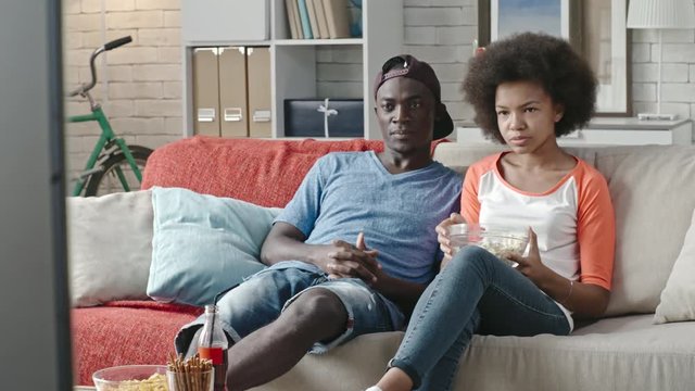 African brother and sister eating popcorn while watching movie on TV at home and discussing something