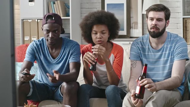 African girl sitting with two friends on couch while watching movie on TV at home. They drinking coke, beer and talking