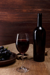 Red wine. Bottle with wineglass. Dark grapes branch on plate. On rustic wooden background.