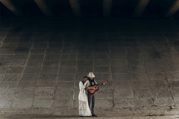 stylish hipster couple hugging under bridge. man in hat playing guitar for his boho woman in...