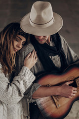 stylish hipster couple gently hugging. man in hat playing guitar for his boho woman in knitted sweater. atmospheric sensual moment. rustic fashionable look.
