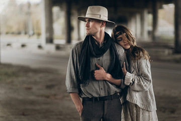 stylish hipster couple gently hugging. boho woman touching arm of confident man in hat under abandoned bridge. atmospheric sensual moment. rustic fashionable look.