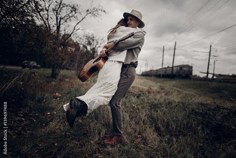 Wall mural man with guitar dancing with his boho gypsy woman in windy field. stylish hipster couple hugging. at