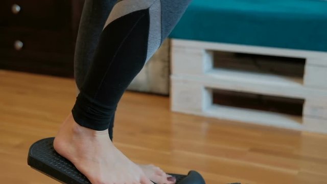 The room woman performs an exercise for the legs on the mini stepper. Sportswoman at home engaged in a compact home cardio equipment. Lady in sports stretch pants stepping on the simulator walk up the