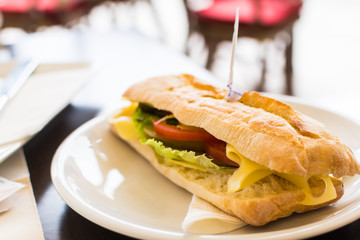 Tasty skewer pierced long ciabatta sandwich with lettuce, slices of fresh tomatoes and cheese on white plate in cafe.