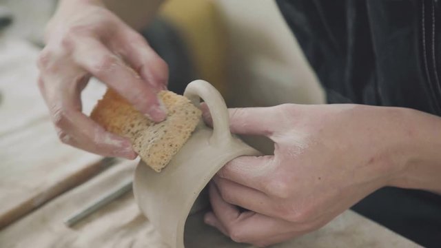 The hands of the potter: a woman ceramist dub blank stein using foam sponge. Close-up of pottery. Clay potter creating in her workshop. Craftsman making pottery. Handmade pottery.