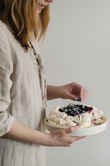 Woman tasting cake. Homemade meringue cake Pavlova with whipped cream, sugar powder, fresh blueberries and blueberry sauce in female hands over gray wall as background, day light.