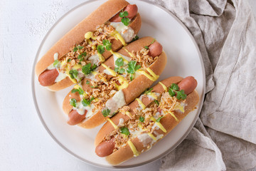 Hot dogs with sausage, fried onion, coriander leaves, cheese sauce and mustard, served on white ceramic plate with textile over white concrete texture background. Fast food. Top view, copy space