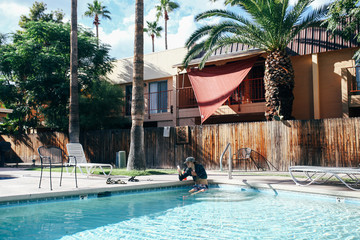 Young trendy man sitting in the pool while looking at his phone surrounded by palm trees