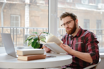 Concentrated bearded young man reading books while using laptop