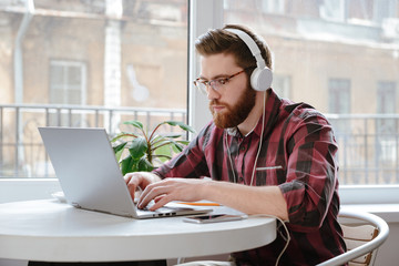 Handsome serious bearded young man using laptop and listening music.