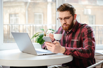 Bearded young man drinking coffee while using laptop and phone.
