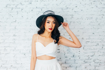 Fototapeta premium Young happy Asian woman smiling in fashionable dress and summer hat over white brick wall