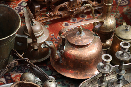 Old things./On an old carpet there are different old metal things. In the centre there is a copper teapot.