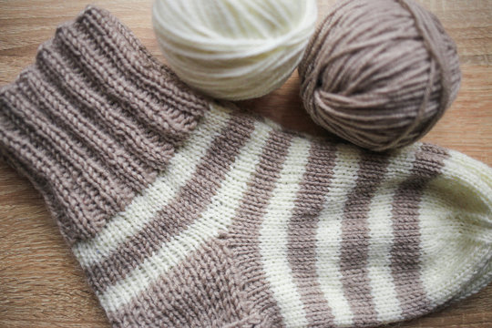 Beige and white yarn, Knitted striped beige-beige sock are on the table. Wooden background. Hobbies 